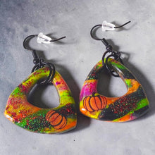 Load image into Gallery viewer, Open-center triangle dangle earrings with pumpkin details, bright glittery Halloween colors, and black ear wires on a white plate